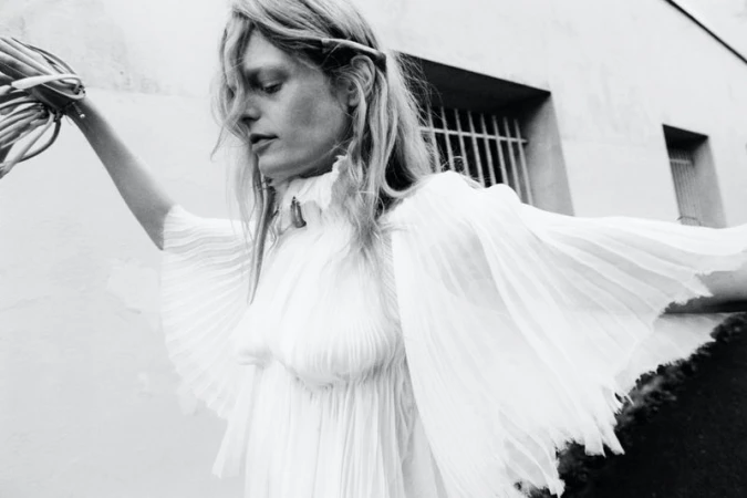 Monochrome fashion portrait of a blonde woman in a flowing white dress, holding foliage, against a textured white wall with barred windows.