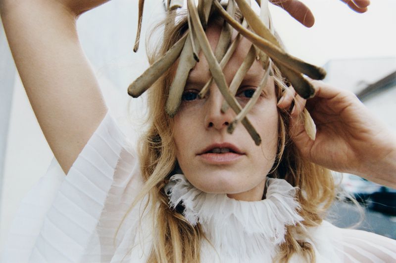 Close-up of a fair-skinned woman wearing a white ruffled collar with light hair and natural elements framing her face, outdoors.