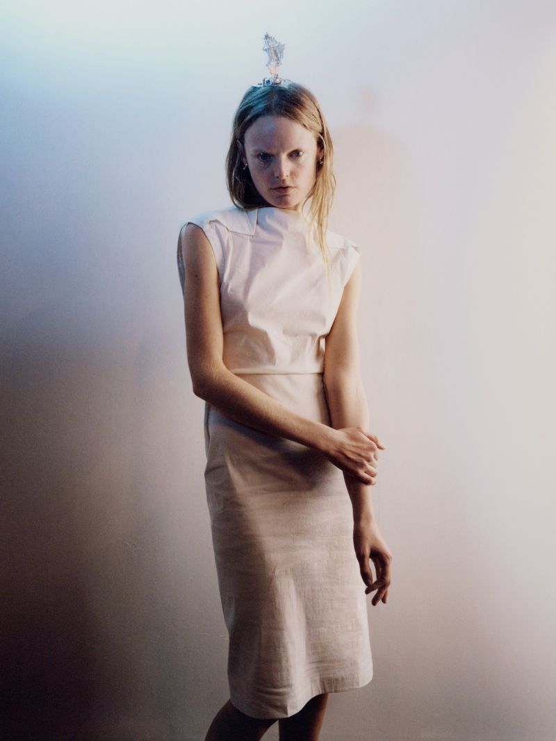 Woman in dress standing in front of gradient background, with sculptural headpiece. Fashion, beauty, studio lighting, neutral tones.