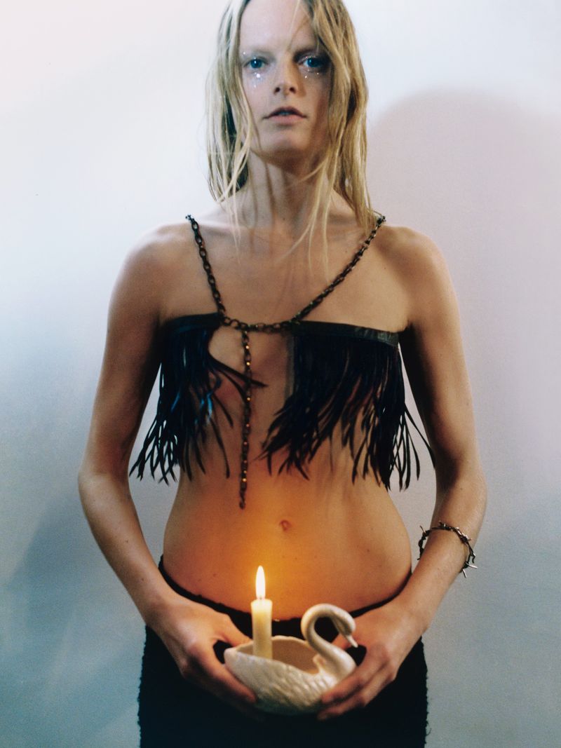 Blonde woman with glitter make-up poses with a candle in a swan-shaped holder, wearing a fringed top against a muted white wall.