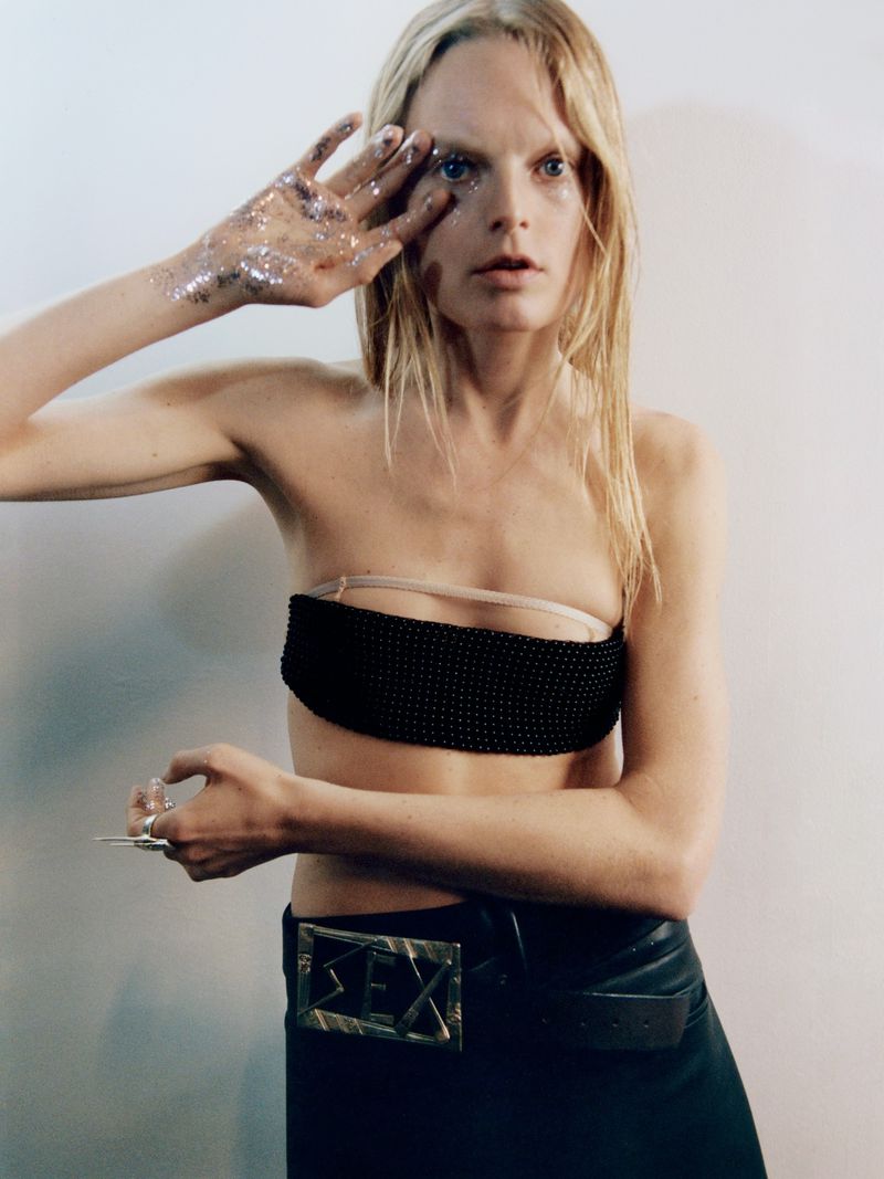 Model with glitter makeup, wearing a black strapless top and a decorative belt, poses against a white wall with a neutral expression.
