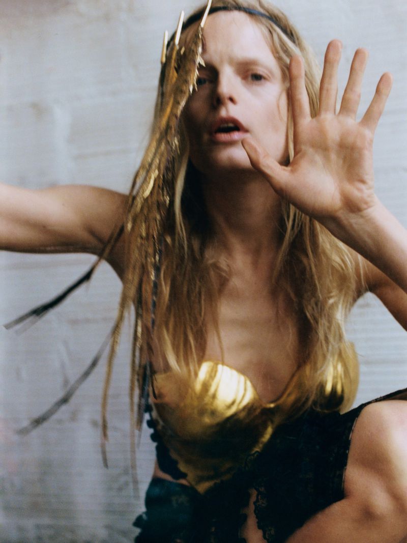Close-up of a fair-skinned woman in a golden bustier, adorned with feathers, partially blocking the view with her hand.