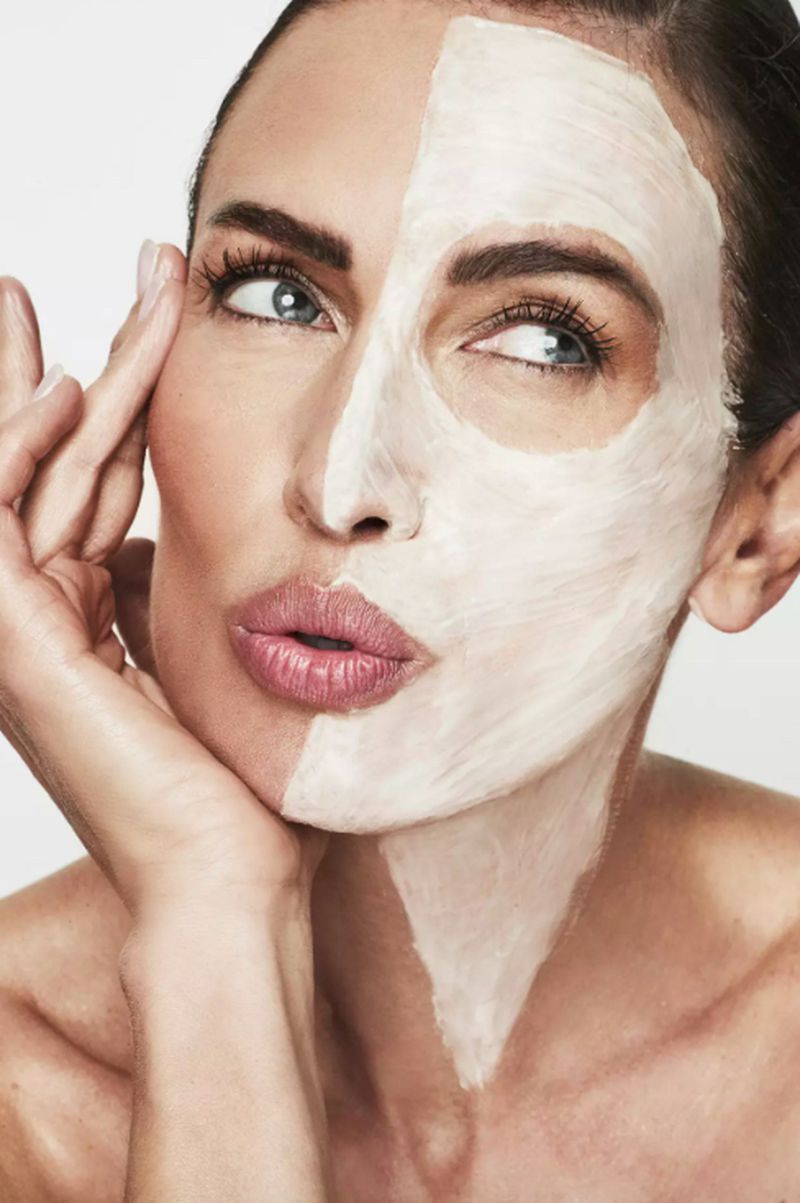 Close-up portrait of a fair-skinned woman with a cosmetic face mask applied to half of her face, looking off to the side.