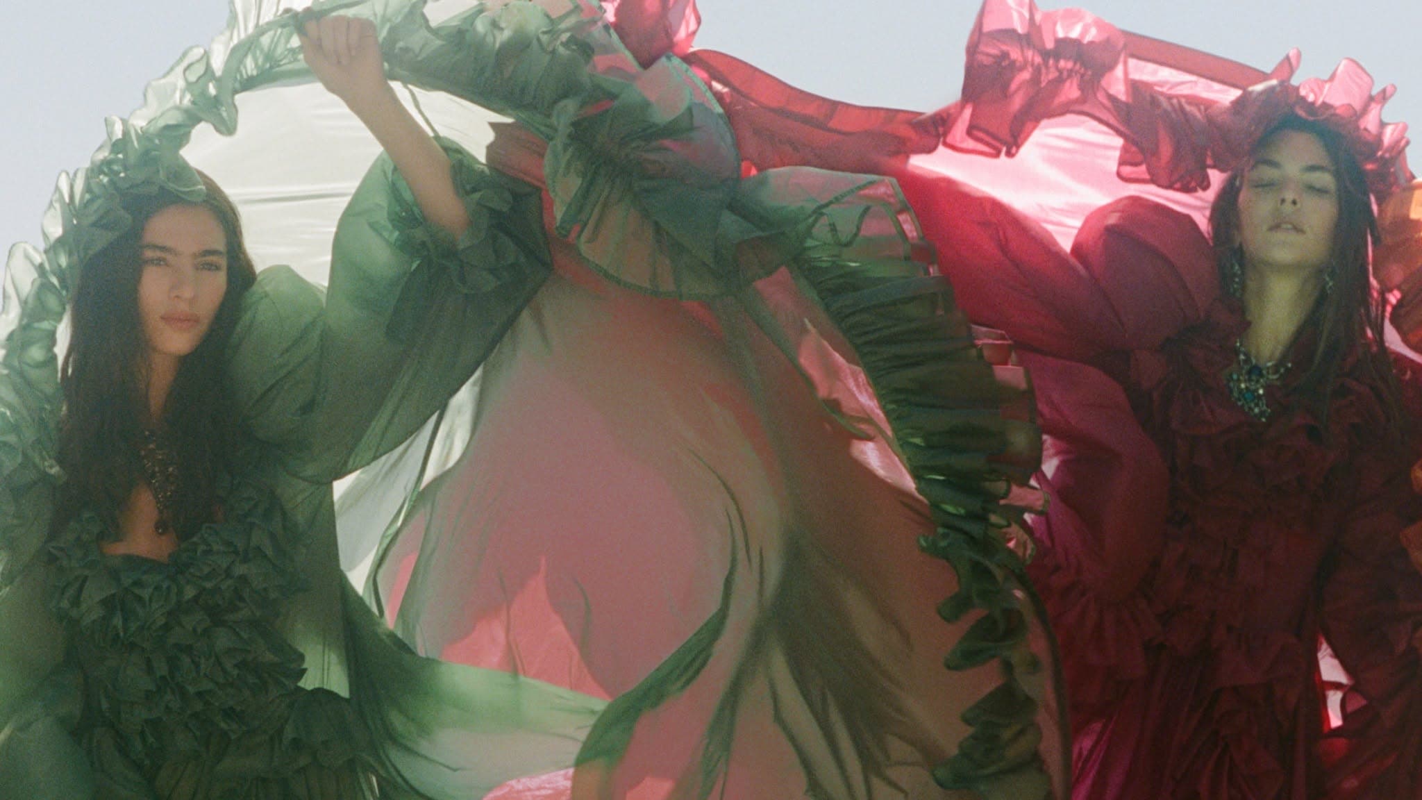 Two models posing in flowing green and crimson sheer ruffled dresses outdoors under soft daylight.