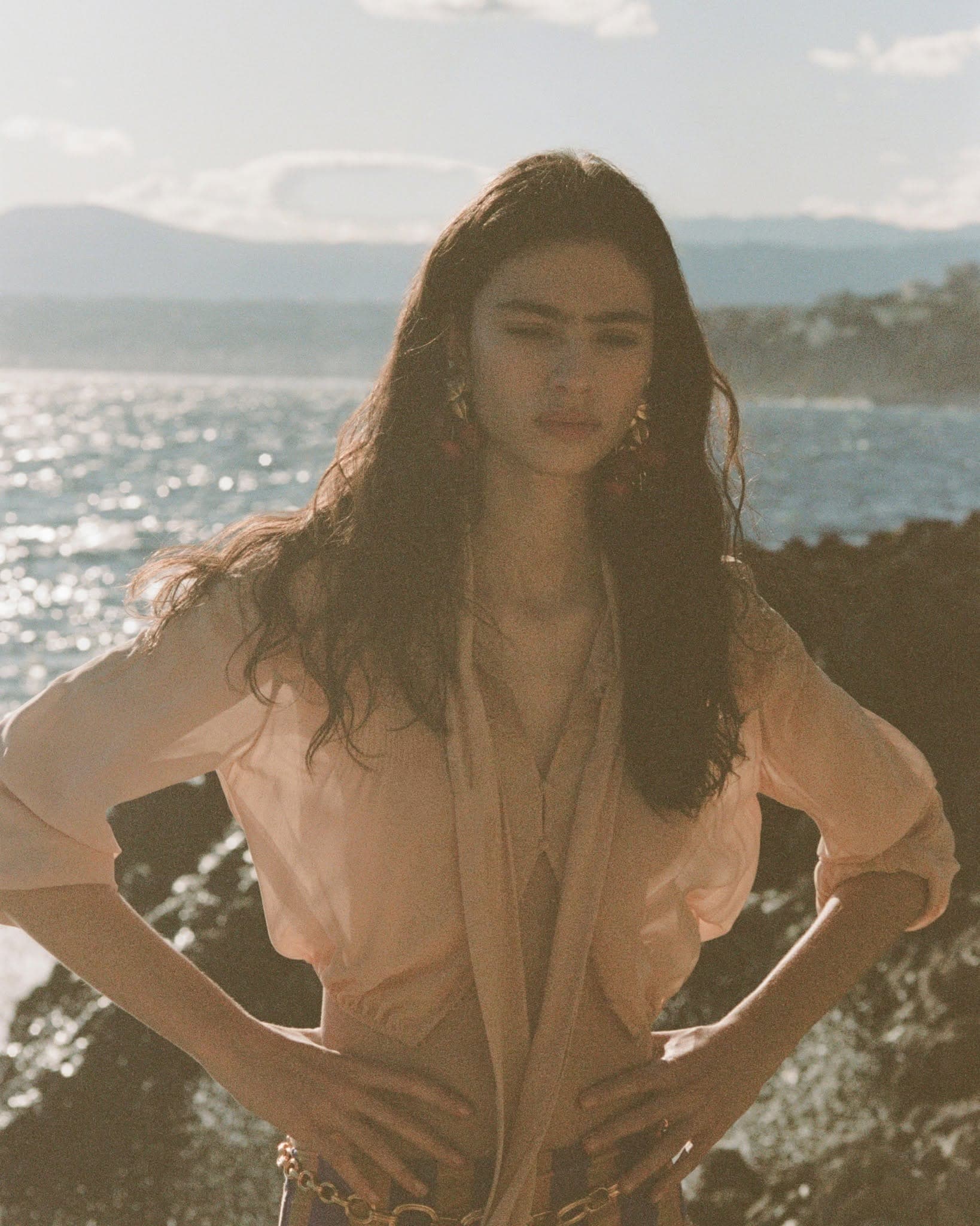 Model posing with hands on hips in a sheer peach blouse against a sunlit rocky coastline and ocean backdrop.
