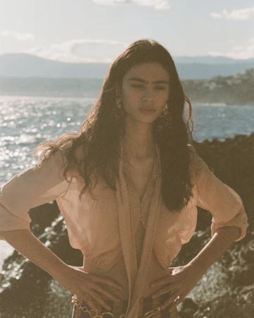 Model posing with hands on hips in a sheer peach blouse against a sunlit rocky coastline and ocean backdrop.