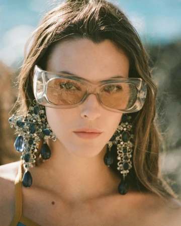 Close-up portrait of a woman wearing chunky clear sunglasses and large ornate crystal earrings outdoors