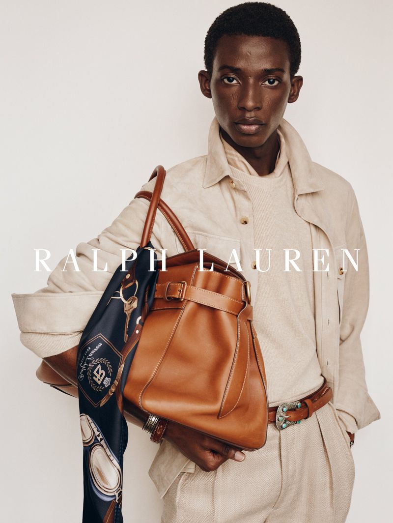 Model Gideon Adeniyi holding a tan leather tote bag with a printed scarf, wearing a beige linen shirt against a white backdrop.