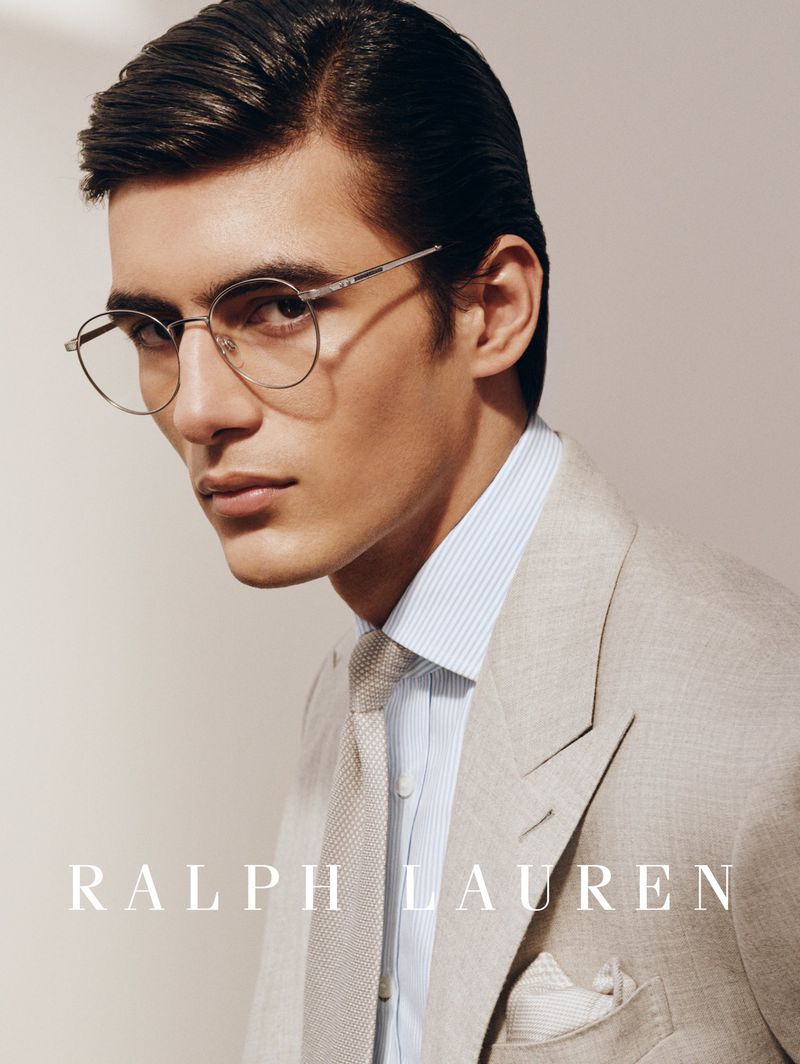 Model wearing round wire-frame eyeglasses, a beige tailored suit jacket, light blue striped shirt, and textured knit tie.