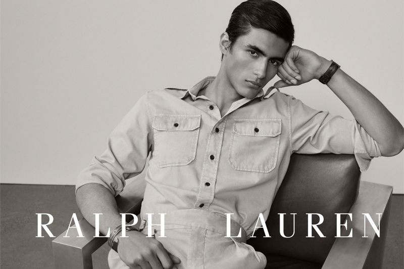 A male model seated in a modern armchair resting his head on his hand, wearing a button-up utility shirt in black and white.