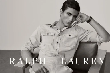A male model seated in a modern armchair resting his head on his hand, wearing a button-up utility shirt in black and white.
