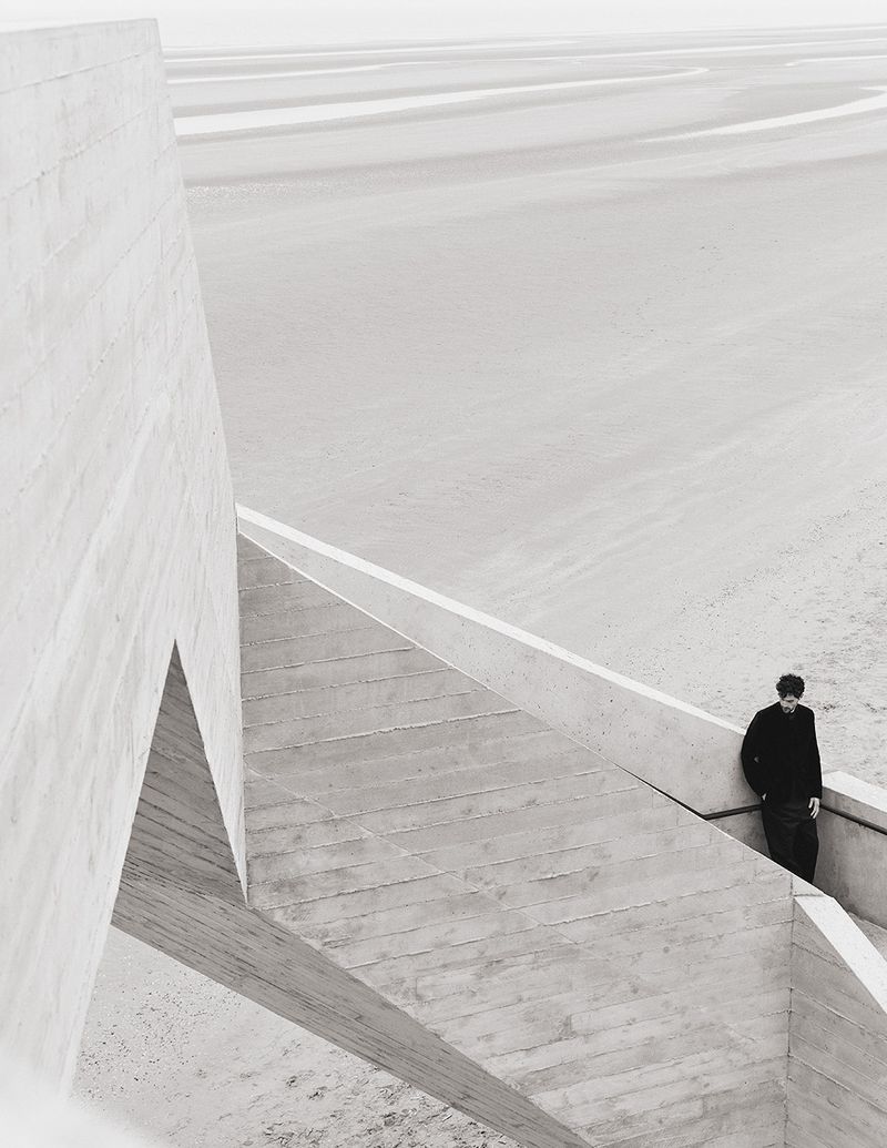 Monochromatic architecture shot of a man standing on concrete stairs next to a modern building structure, with an open sandy landscape.