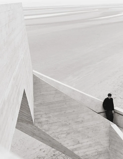 Monochromatic architecture shot of a man standing on concrete stairs next to a modern building structure, with an open sandy landscape.