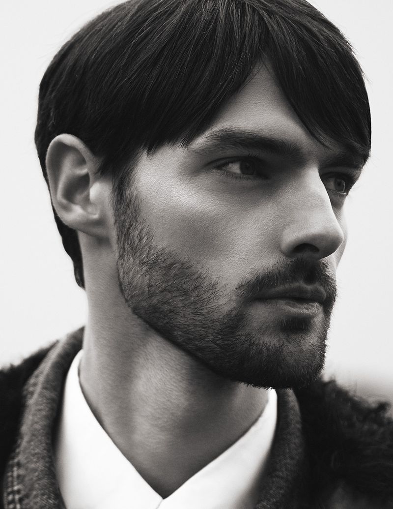 Close-up portrait of a bearded man with dark hair wearing a collared shirt and jacket, gazing to the side against a neutral background.