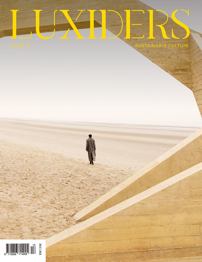 A person walks along a beige desert landscape towards the horizon, framed by modern concrete architectural elements, magazine cover.