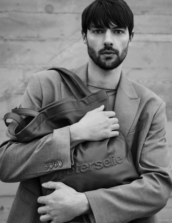 Monochrome portrait of a male model in a blazer holding a large tote bag against a textured concrete background.