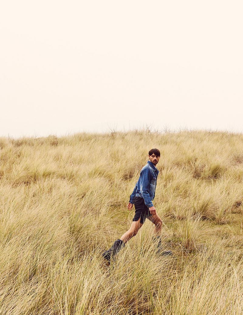 Man in denim jacket walks through a field of tall yellow grass under an overcast sky, looking at the camera in a medium shot.