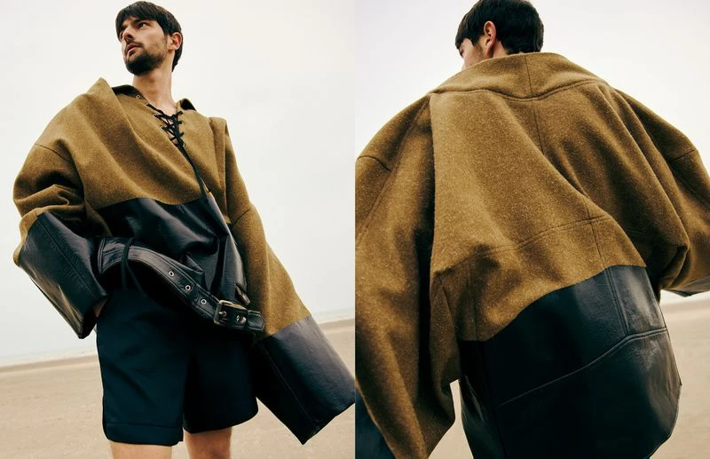 Fashion shot: A man with a beard wearing a large olive coat with black leather accents and black shorts on a sandy beach backdrop.
