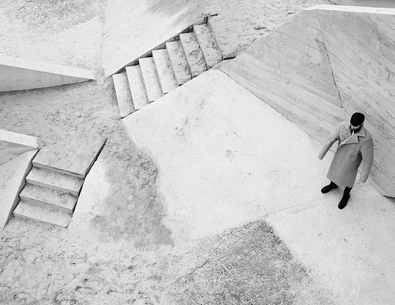 Monochrome portrait of a man in an overcoat standing on a concrete structure with stairs, creating an abstract architectural view.