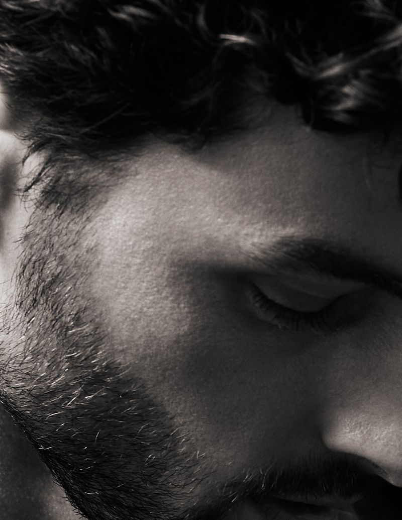 Monochrome close-up of a bearded man's face featuring his eyes closed, and focusing on his textured beard and curly dark hair.