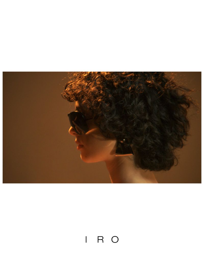 Model with dark curly hair and sunglasses shown in profile against a warm backdrop with IRO logo below.