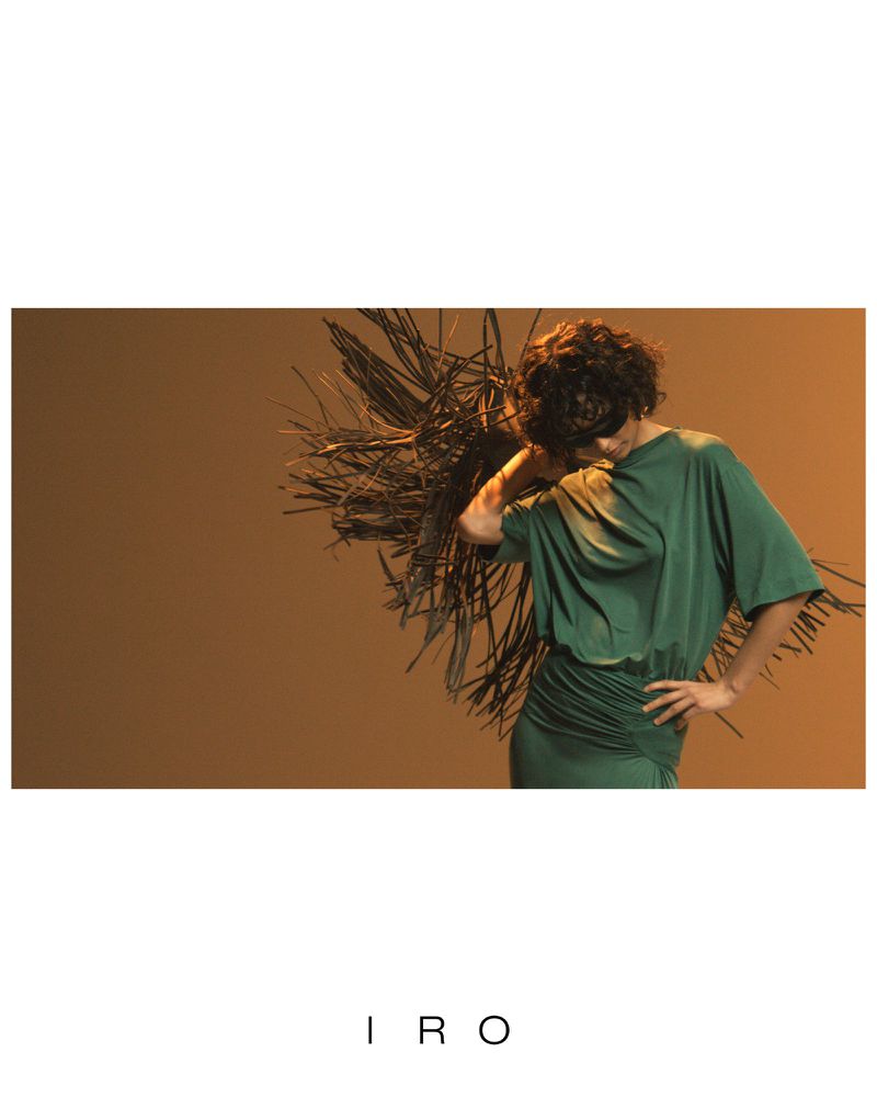 Model with short dark curly hair in a draped green dress posing against a warm brown background with dry branch textures for IRO.