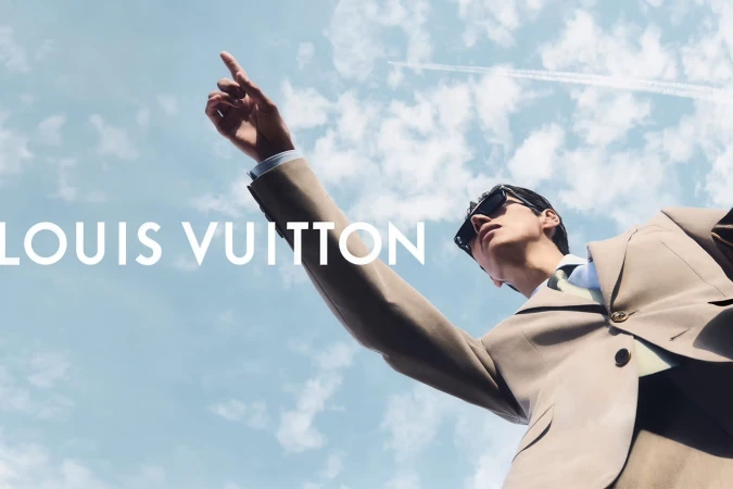 Low-angle shot of a man in a beige tailored suit and sunglasses pointing upward toward a blue sky.