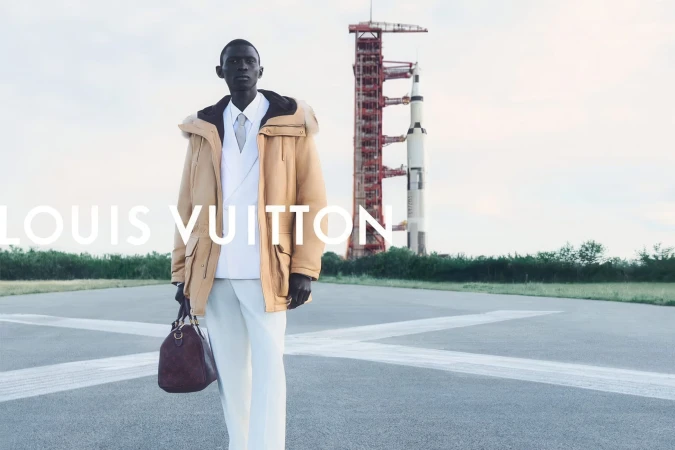 Black male model in a camel parka and white suit holding a brown leather duffle bag on a tarmac runway with a rocket behind him.