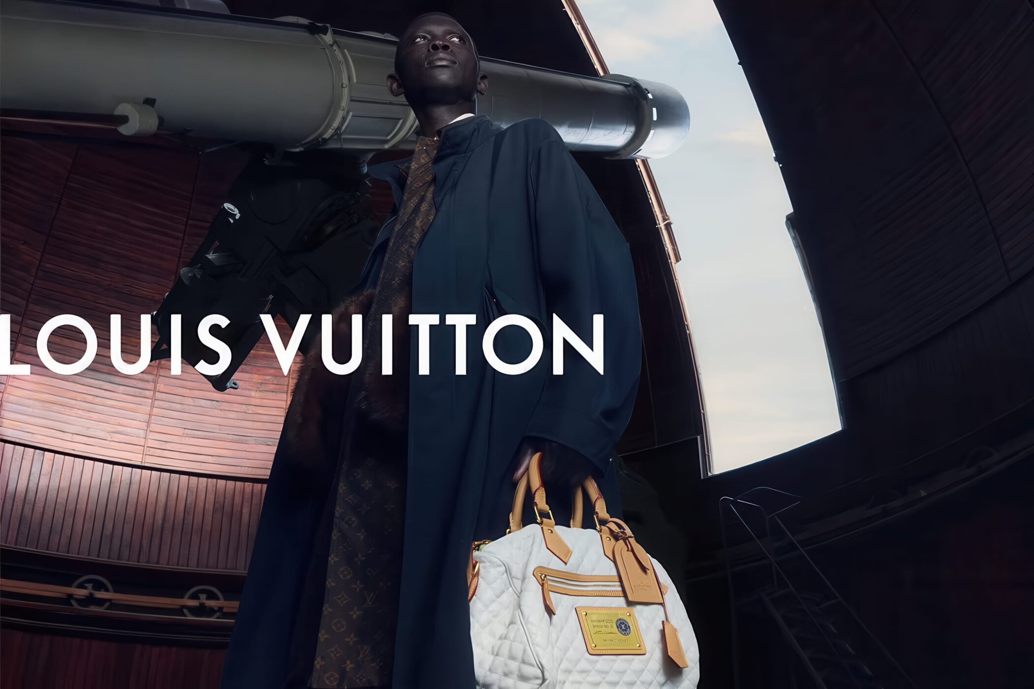 Black male model wearing a dark coat and holding a white quilted Louis Vuitton handbag in an observatory beside a large telescope.