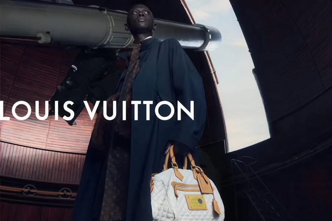 Black male model wearing a dark coat and holding a white quilted Louis Vuitton handbag in an observatory beside a large telescope.