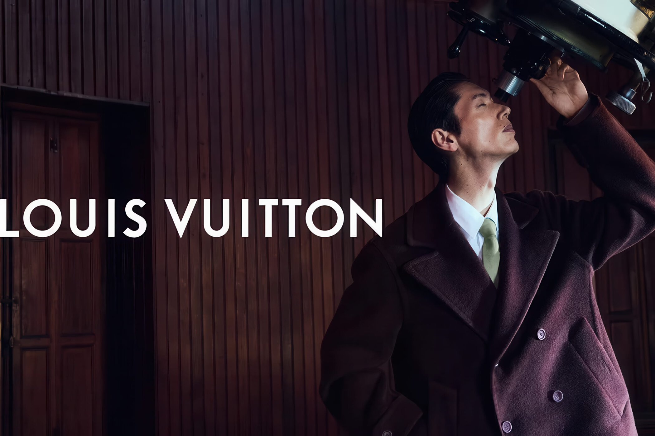 Man in a dark burgundy wool coat and tie looking through an astronomical telescope in a wood-paneled room.