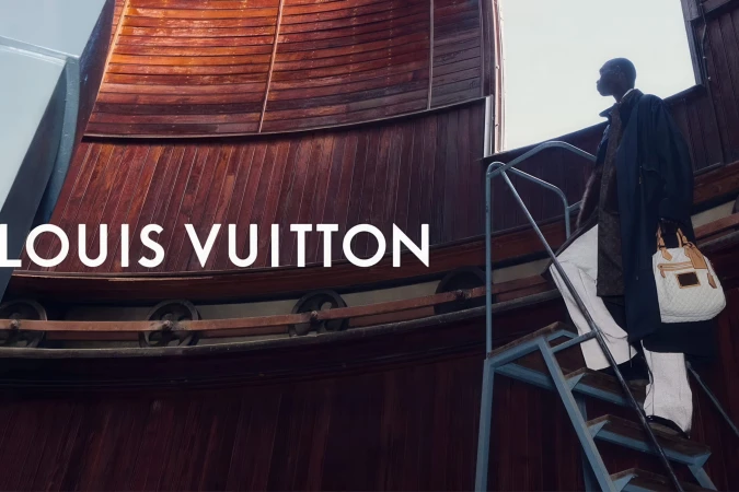 Model carrying a white quilted tote bag ascending metal stairs against a curved wooden architectural wall with Louis Vuitton logo text.