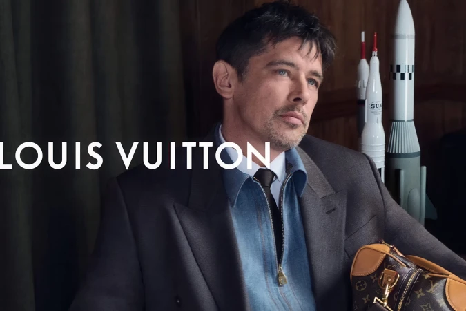 Man wearing a tailored suit jacket over a blue denim shirt holding a monogram duffle bag with miniature rocket models in the background.