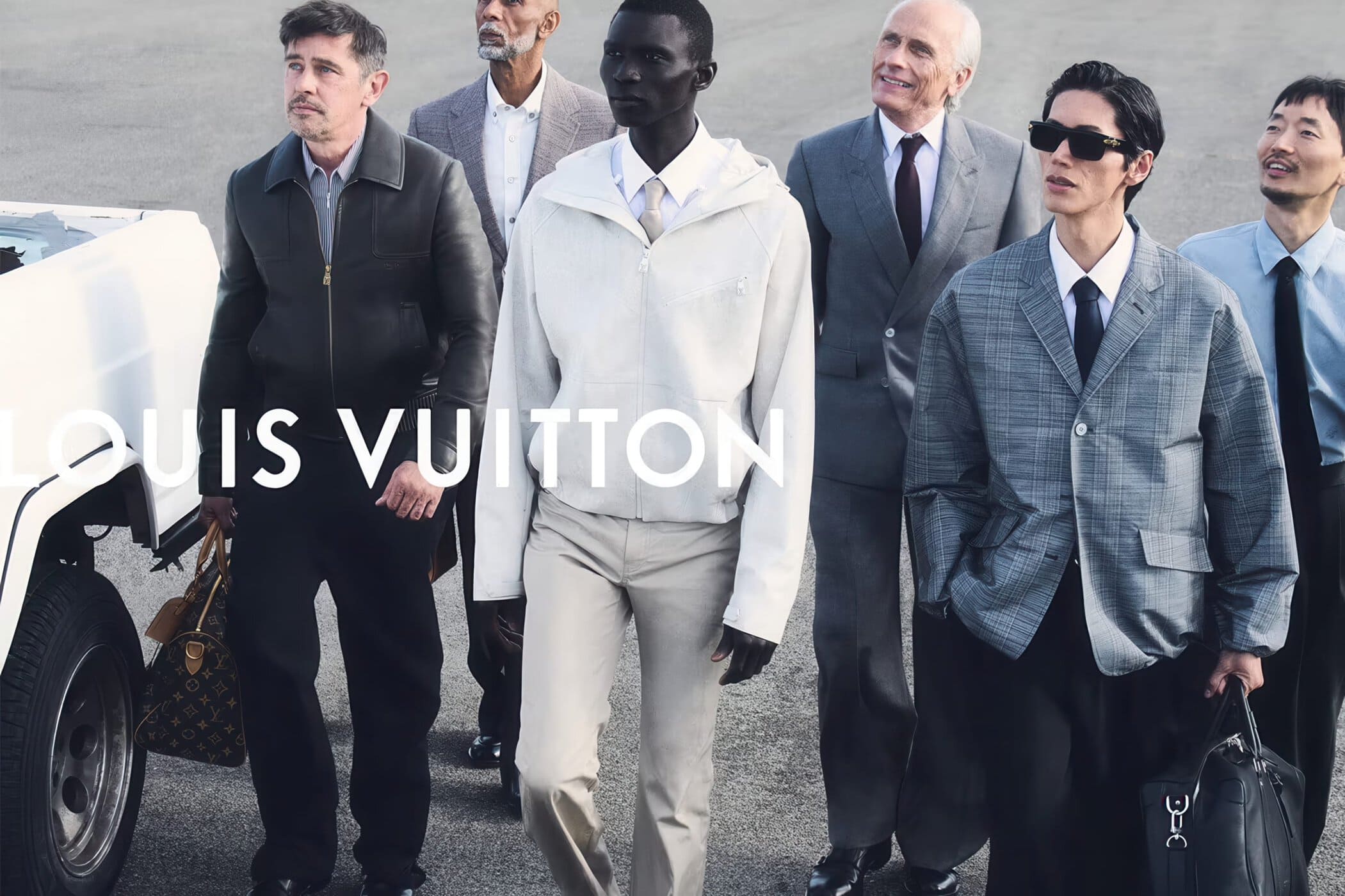 Group of diverse male models in tailored Louis Vuitton menswear walking together on tarmac next to a white truck.