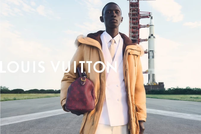 Model standing on a tarmac holding a burgundy monogram handbag in front of a rocket launch structure for Louis Vuitton.