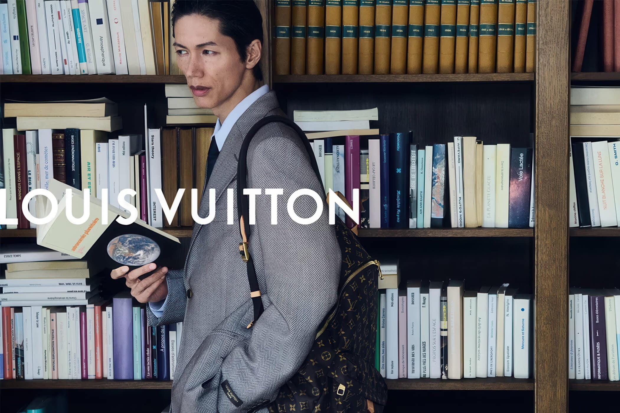 An Asian man in a tailored grey blazer holds an open book while carrying a monogram backpack in front of a wooden library bookshelf.