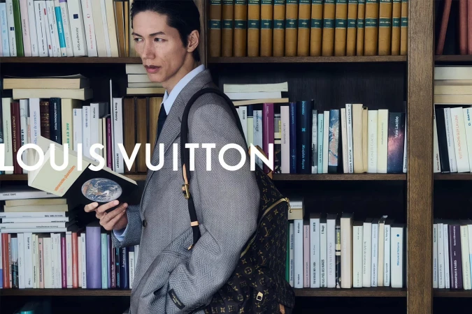 An Asian man in a tailored grey blazer holds an open book while carrying a monogram backpack in front of a wooden library bookshelf.