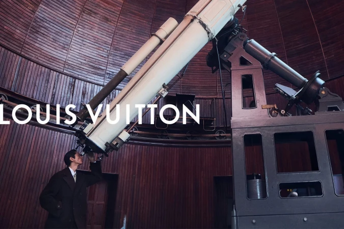 A man in a dark tailored overcoat peers through a massive vintage telescope inside a wooden observatory dome.