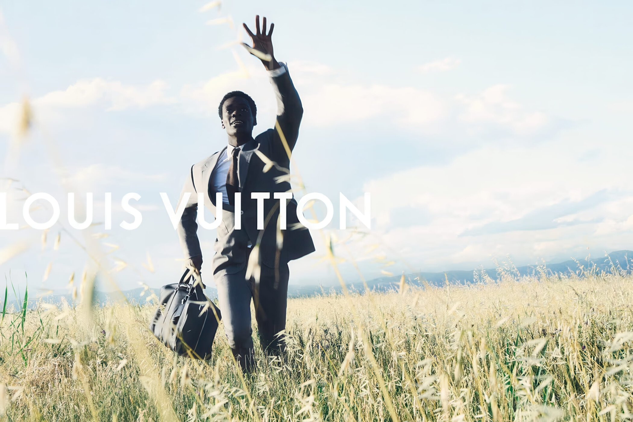 Man wearing a suit waving while walking through a tall grass field holding a dark travel bag