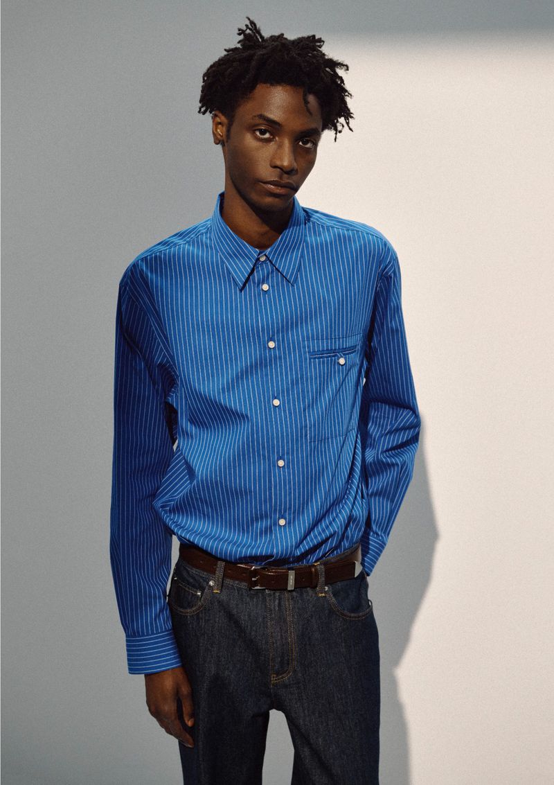 Ahmed Richards wears a blue striped button-up shirt and dark denim jeans with a brown leather belt against a neutral studio backdrop.