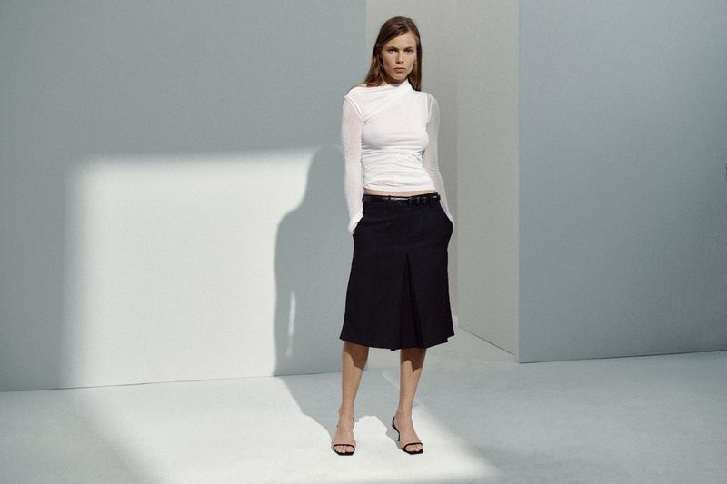 Model wearing a white ruched long-sleeve top and black pleated knee-length skirt in a minimalist grey studio.