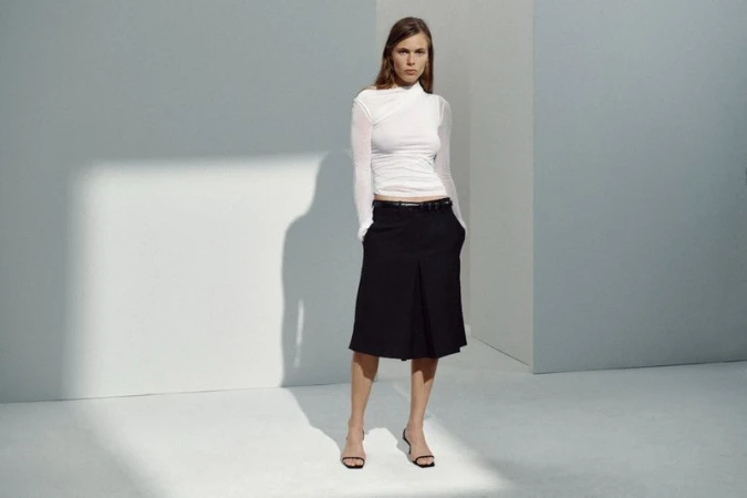 Model wearing a white ruched long-sleeve top and black pleated knee-length skirt in a minimalist grey studio.