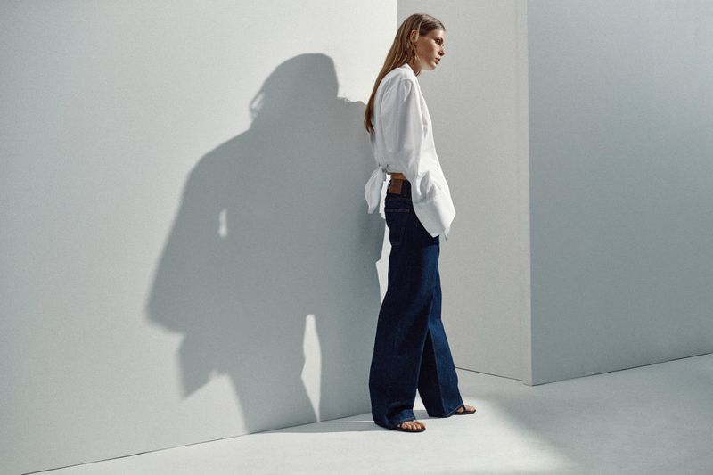Model wearing a white shirt and wide-leg dark denim jeans casting a defined shadow against a minimalist studio wall.