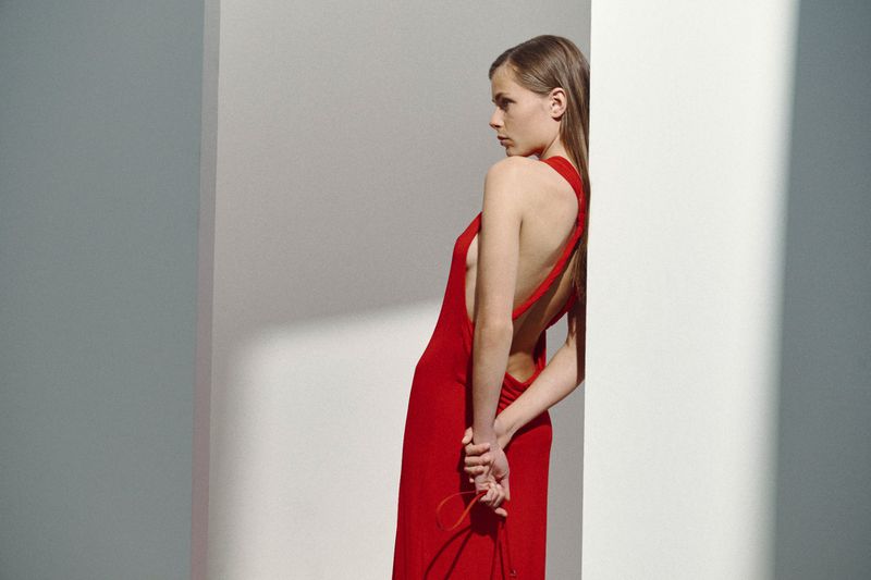 A woman in a vibrant red backless halter dress leans against a white architectural column in profile under angled light.
