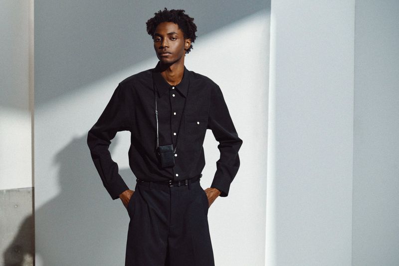 Model Ahmed Richards wearing a black button-up shirt and tailored trousers with a small neck pouch against a white wall.
