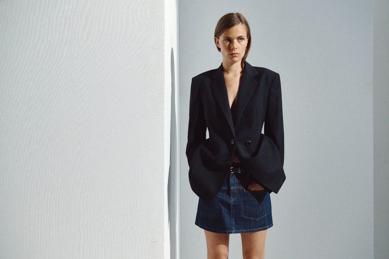Model wearing an oversized structured black blazer and denim miniskirt standing against a white studio wall.
