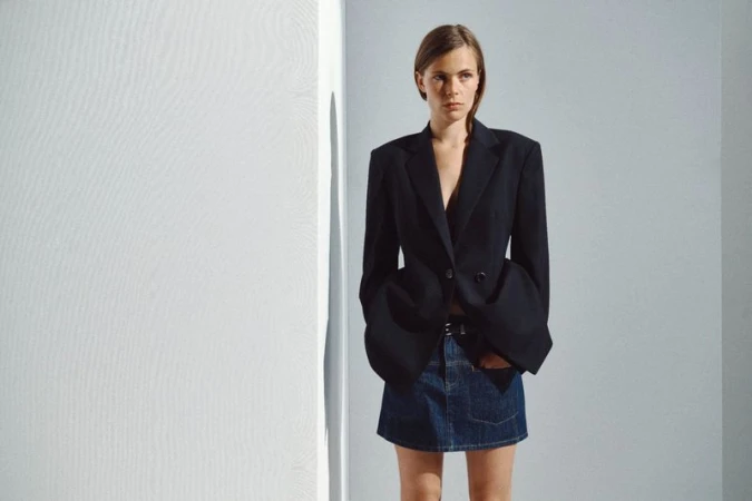 Model wearing an oversized structured black blazer and denim miniskirt standing against a white studio wall.