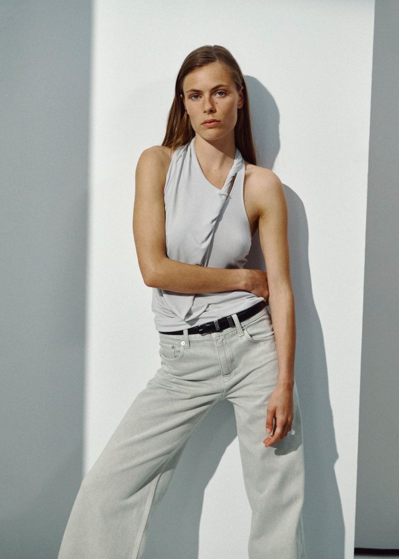 Model wearing a pale draped sleeveless top and light wash denim jeans posing against a white studio wall.