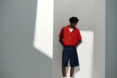 Model Ahmed Richards in a bright red short-sleeve shirt and dark denim shorts standing against a grey wall in sharp geometric sunlight.