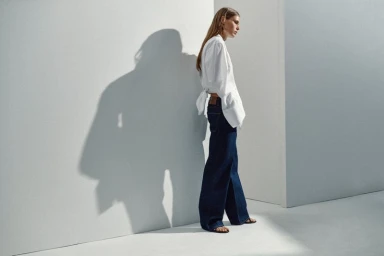 Model wearing a white shirt and wide-leg dark denim jeans casting a defined shadow against a minimalist studio wall.